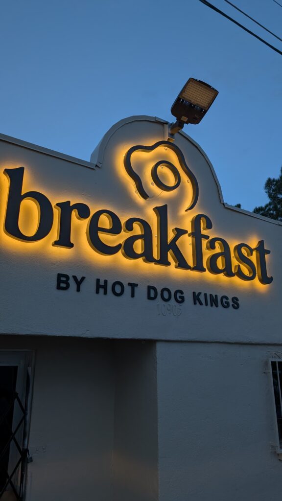 Fachada Breakfast by Hot Dog Kings con letrero cursiva dorada retroiluminada y huevo icono de noche