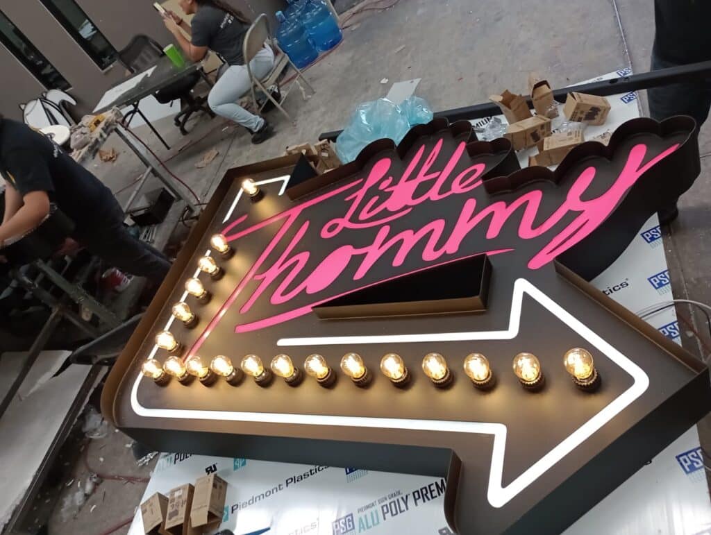 Letrero flecha Little Thommy con script rosa neon y bombillas doradas en contorno