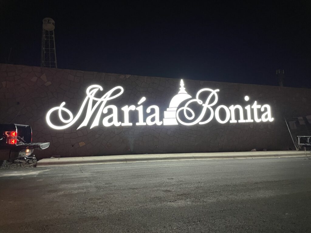 Letrero María Bonita en script blanco sobre pared gris de noche