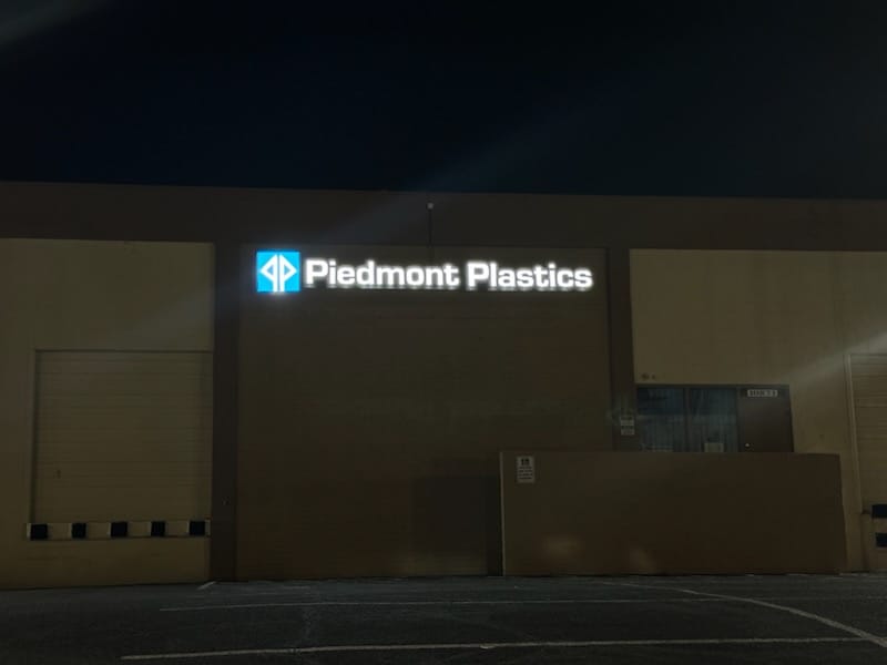 Letrero Piedmont Plastics blanco retroiluminado con logo rombo azul de noche
