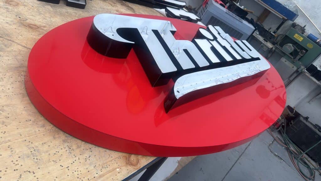 Letrero Thrifty script plateado sobre base circular roja cableado LED visible