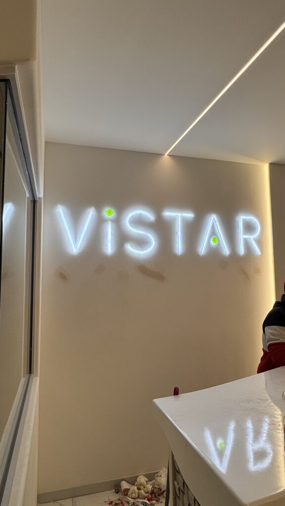 Letrero neon turquesa Vistar con detalles en verde sobre pared beige interior